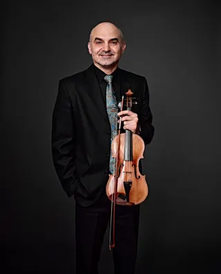 Adrian Petrutiu is a bald white man. He is wearing a black shirt and suit jacket, a blue and gold tie, and holding his violin.