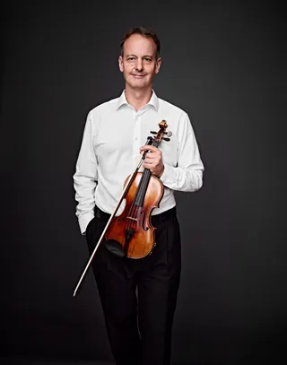 Andreas Uhlemann is a white man with gray hair. He is wearing a white shirt, smiling, and holding a violin.