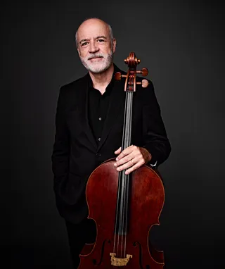 Bráulio Lima is a bald white man with a white beard. He is wearing a black shirt and suit jacket, and he is holding a cello.