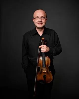 Cristian Sandu is a bald white man wearing glasses. He is dressed in a black shirt and holding a violin.