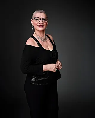 Cristiane Minczuk é uma mulher branca, com a cabeça raspada e usando óculos. Ela está usando brincos longos, um colar prateado e uma roupa preta, e está sorrindo.