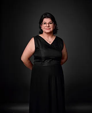Eliane Chagas é uma mulher branca, com cabelo preto na altura dos ombros. Ela usa óculos redondos de armação fina e um vestido preto sem mangas com decote em V. No pescoço, ela usa um colar dourado.