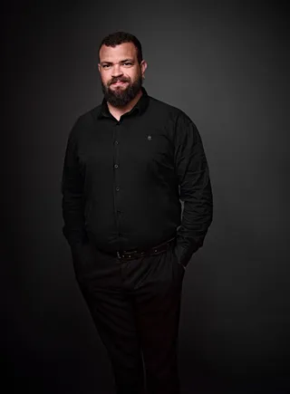 Erick Souza é um homem branco, adulto, com cabelo preto curto e barba longa. Ele está usando uma camisa preta e tem um brinco na orelha esquerda.