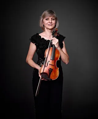 Katia Spassova is a white woman with short blonde hair. She is wearing a black dress with ruffles and holding a violin.