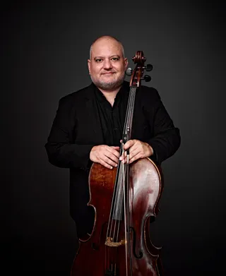 Rodrigo Andrade é um homem branco, calvo. Ele está usando roupas pretas e segurando um violoncelo.