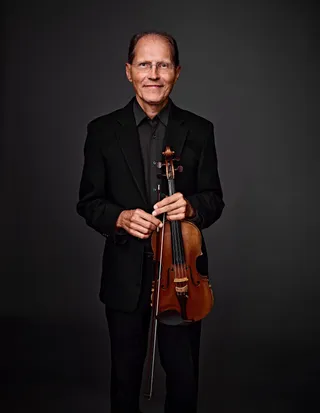 Yuriy Rakevich é um homem branco, calvo, de olhos claros. Ele usa óculos, camisa e paletó pretos.