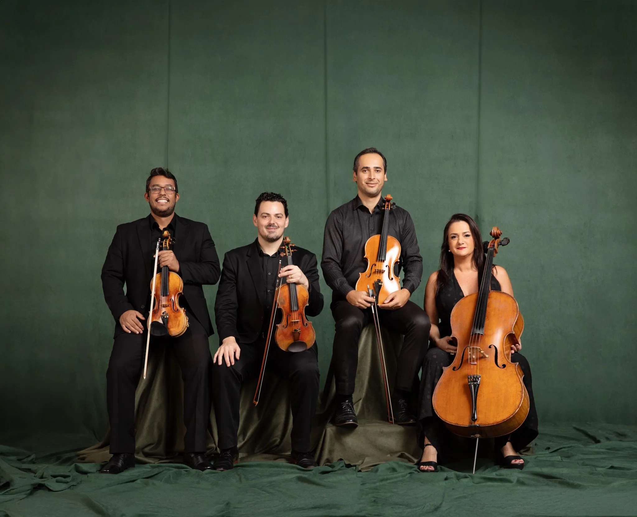 O quarteto de cordas está sentado com seus instrumentos no colo. Eles usam preto e sorriem à frente de um fundo verde.
