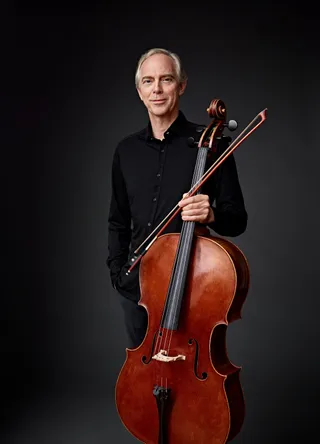 Kim Bak Dinitzen é um homem branco, grisalho e de olhos azuis. Ele veste preto e segura um violoncelo.