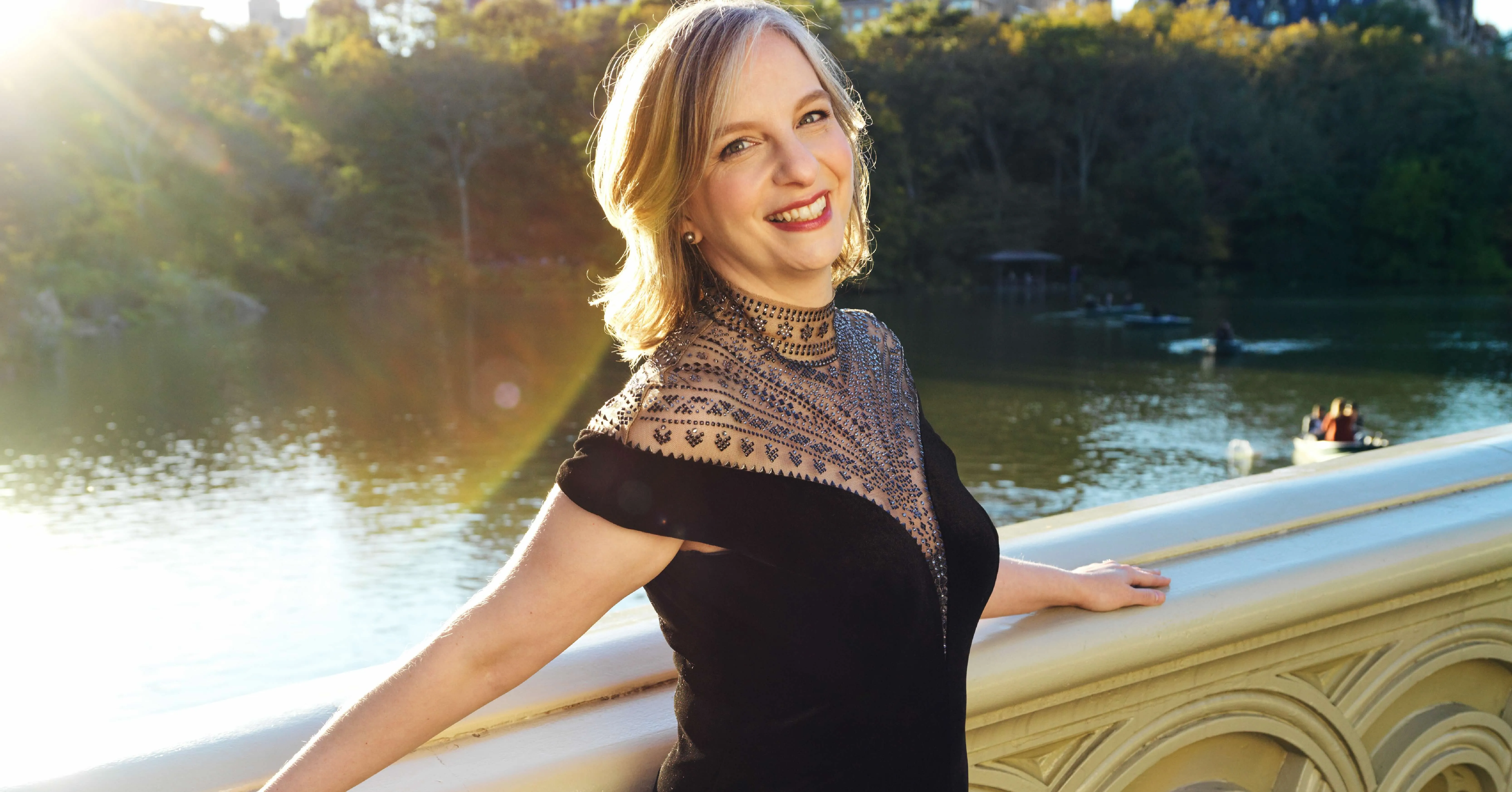 Orli Shaham é uma mulher branca, loira. Ela sorri apoiada em uma ponte, com um rio ensolarado atrás.