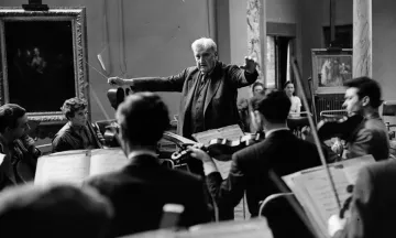 Foto em preto e branco de Ralph Vaughan Williams, homem branco e idoso, com cabelos brancos. Ele está de pé, com braços abertos e batuta na mão, regendo um grupo de músicos sentados a sua frente.