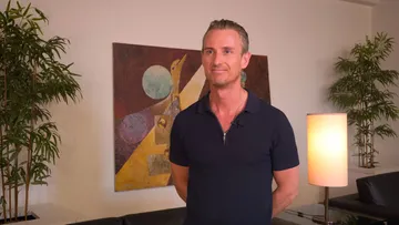 Foto de Alexander Shelley, um homem branco, de cabelos curtos e grisalhos. Ele usa camisa social preta de gola em formato v. Atrás do maestro, pode se ver um quadro vermelho grande, duas plantas altas verdes e um abajur aceso.