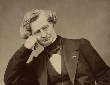 Retrato em sépia de Hector Berlioz, com expressão pensativa e rosto apoiado na mão. Usa traje formal com gravata borboleta, paletó escuro e colete, em frente a fundo neutro que destaca seu semblante melancólico.