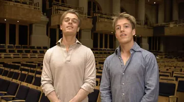 Foto de Arthur Jussen e Lucas Jussen. Arthur está na esquerda, é um homem branco, loiro e veste uma camiseta bege com golas abertas. Lucas está na direita, é um homem branco, de cabelos loiros e veste uma camisa azul de botões.