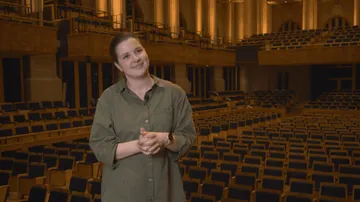 Emilia é uma mulher branca de cabelo castanho preso, camisa verde-oliva, está em pé no palco de uma sala de concertos vazia, sorrindo levemente com as mãos cruzadas à frente do corpo. Ao fundo, vemos fileiras de cadeiras e varandas iluminadas.