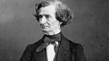 Retrato em preto e branco de Hector Berlioz, com expressão pensativa e olhando para o lado esquerdo. Usa traje formal com gravata borboleta, paletó escuro e colete, em frente a fundo neutro que destaca seu semblante melancólico.