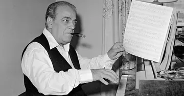 Fotografia em preto e branco de Heitor Villa-Lobos ao piano, com expressão concentrada. Usa camisa branca, colete escuro e fuma um charuto, segurando uma partitura em ambiente interno com luz suave.