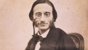 Retrato de Jacques Offenbach, compositor francês do século XIX, com expressão serena, usando óculos de monóculo, costeletas marcantes e traje formal escuro, sentado diante de fundo neutro.