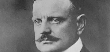 Retrato em preto e branco de Jean Sibelius, compositor finlandês, com olhar sério e bigode espesso, vestindo paletó e camisa de colarinho alto, diante de fundo escuro e neutro.