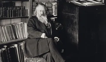 Foto de Johannes Brahms, homem branco, de barbas e cabelos brancos e compridos. Ele está sentado com uma perna cruzada sobre a outra.