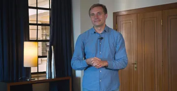 Foto de Vasily Petrenko, homem de meia-idade, vestindo camisa azul e jeans, está em pé em um ambiente interno bem iluminado. Ele sorri levemente para a câmera, com as mãos entrelaçadas à frente do corpo.