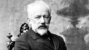 Fotografia em preto e branco de Piotr Ilitch Tchaikovsky com expressão sóbria e olhar direto. Usa paletó escuro com gola alta, sentado em uma cadeira ornamentada, diante de um fundo neutro com padrões suaves e sombreados.