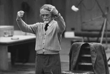 Foto em preto e branco de Zoltán Kodály em ensaio, de pé com os braços erguidos em gesto expressivo de regência. Usa suéter claro, gravata e tem expressão concentrada; ao lado, um paletó repousa sobre uma cadeira.