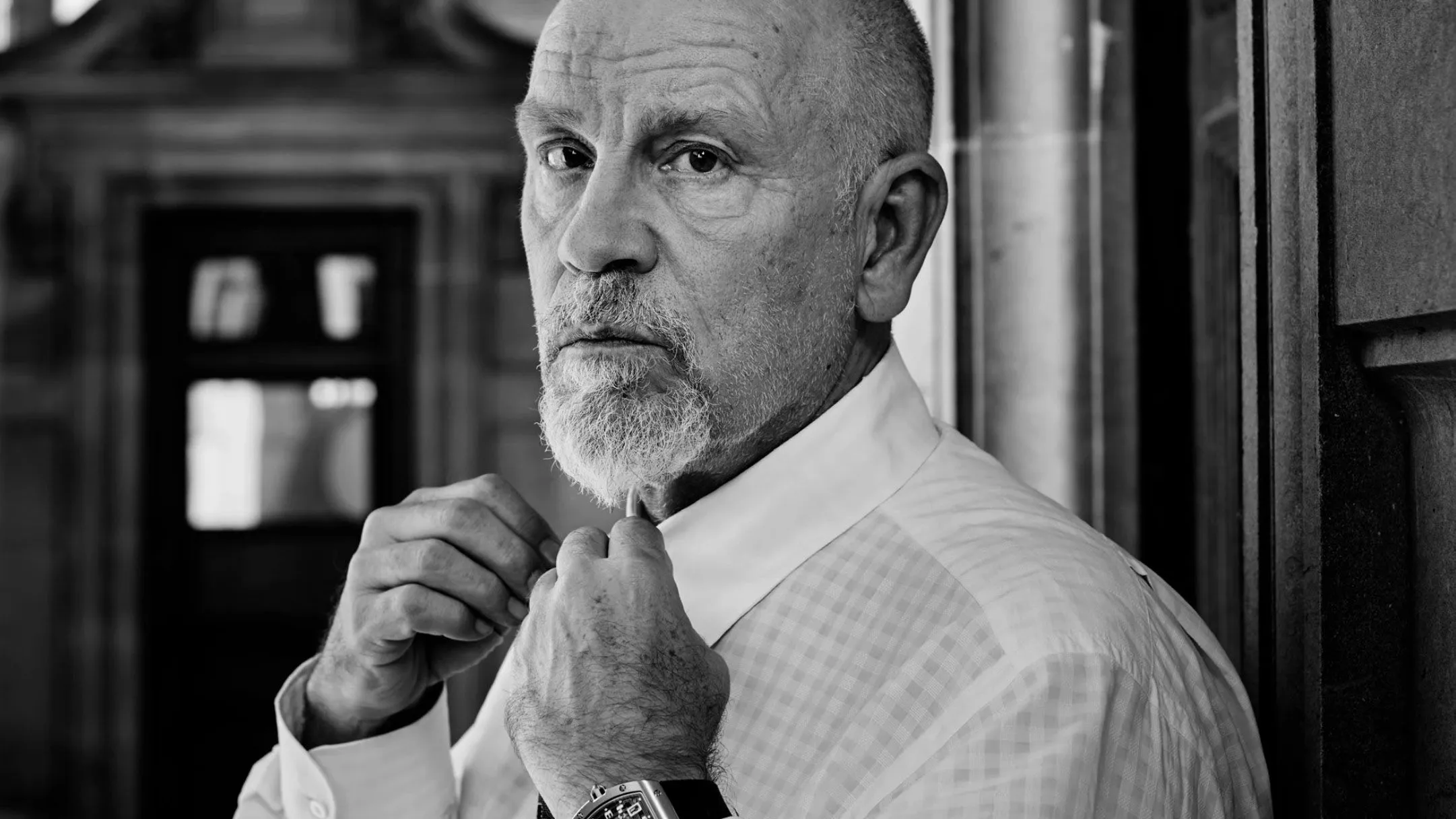 Foto de John Malkovich , homem mais velho, careca, com barba branca curta, ajustando o colarinho da camisa. A foto é em preto e branco, com expressão séria, em um ambiente interno.