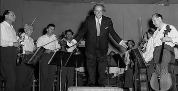 Foto em preto e branco de Heitor Villa-Lobos, homem adulto, branco, em traje formal de calça e blazer escuros, camisa clara de gravata. Ele está de pé sobre um pulpuito e diversos músicos ao seu redor estão levantados em posição de agradecimento.