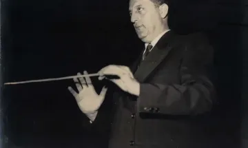 Foto em preto e branco de Francisco Mignone, maestro e compositor brasileiro, com batuta em mãos em gesto de regência. Veste terno escuro e gravata, diante de fundo preto que realça sua figura e expressão concentrada.