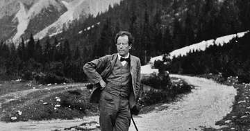 Foto em preto e branco mostra Gustav Mahler em uma estrada de terra, cercado por montanhas e floresta. Ele está de pé, com uma mão na cintura e outra apoiada em uma bengala, usando terno e gravata borboleta.