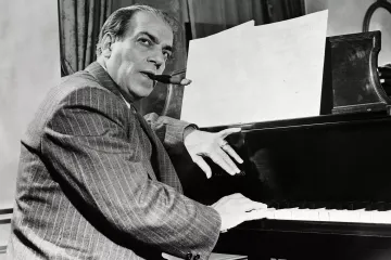 Villa-Lobos é um homem branco, de terno e gravata. Ele está sendado ao lado de um piano e fuma um charuto.