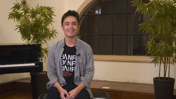 Foto do violoncelista Jay Campbell. Homem adulto, branco e de cabelos escuros e curtos. Ele está sentado em frente a um piano e veste camisa azul clara por cima de camiseta preta. Ele sorri para a foto.