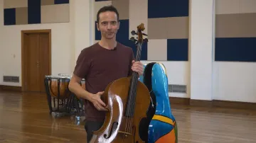 Foto de Jean-Guihen Queyras, homem adulto e branco, de cabelos curtos e pretos. Ele veste uma camiseta marrom e segura, a frente do corpo, seu violoncelo.
