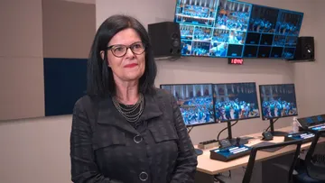 Foto de Yara Caznok, mulher branca, de cabelos pretos e longos. Ela usa óculos pretos e retangulares, blusa preta e colares prateados. No fundo, é possível ver telas do estúdio de gravação da Osesp.